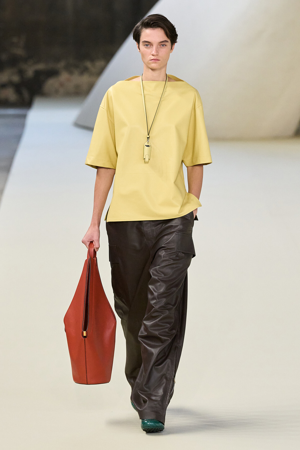 Model walking the runway in a loose pale yellow top with mid-length sleeves, dark brown leather cargo pants, green shoes, and a large red tote bag. Necklace with a pendant visible.