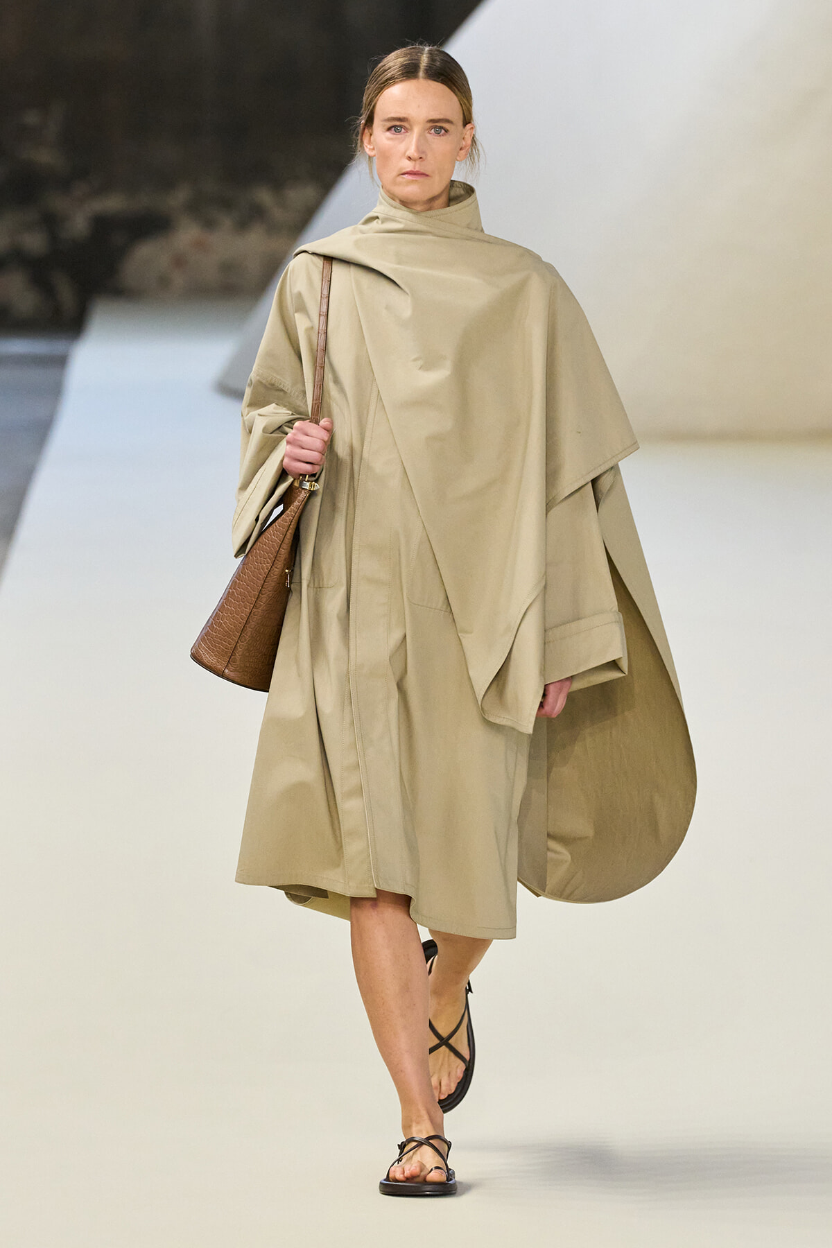 Model walks the runway in a beige oversized cape coat, carrying a brown textured tote and wearing strappy sandals.