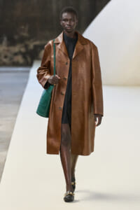 Female model on a runway wearing a long brown leather coat over a black outfit, carrying a green textured handbag.