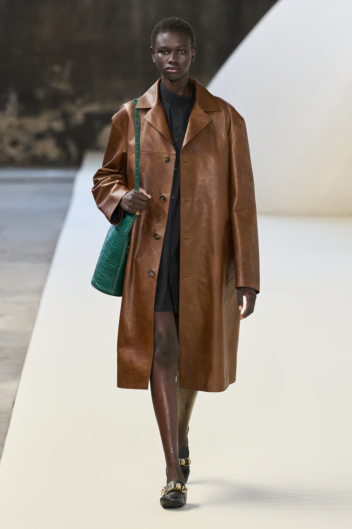 Female model on a runway wearing a long brown leather coat over a black outfit, carrying a green textured handbag.