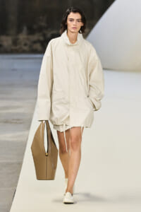 Model on a runway wearing an oversized cream coat with a drawstring hem, carrying a large tan tote.