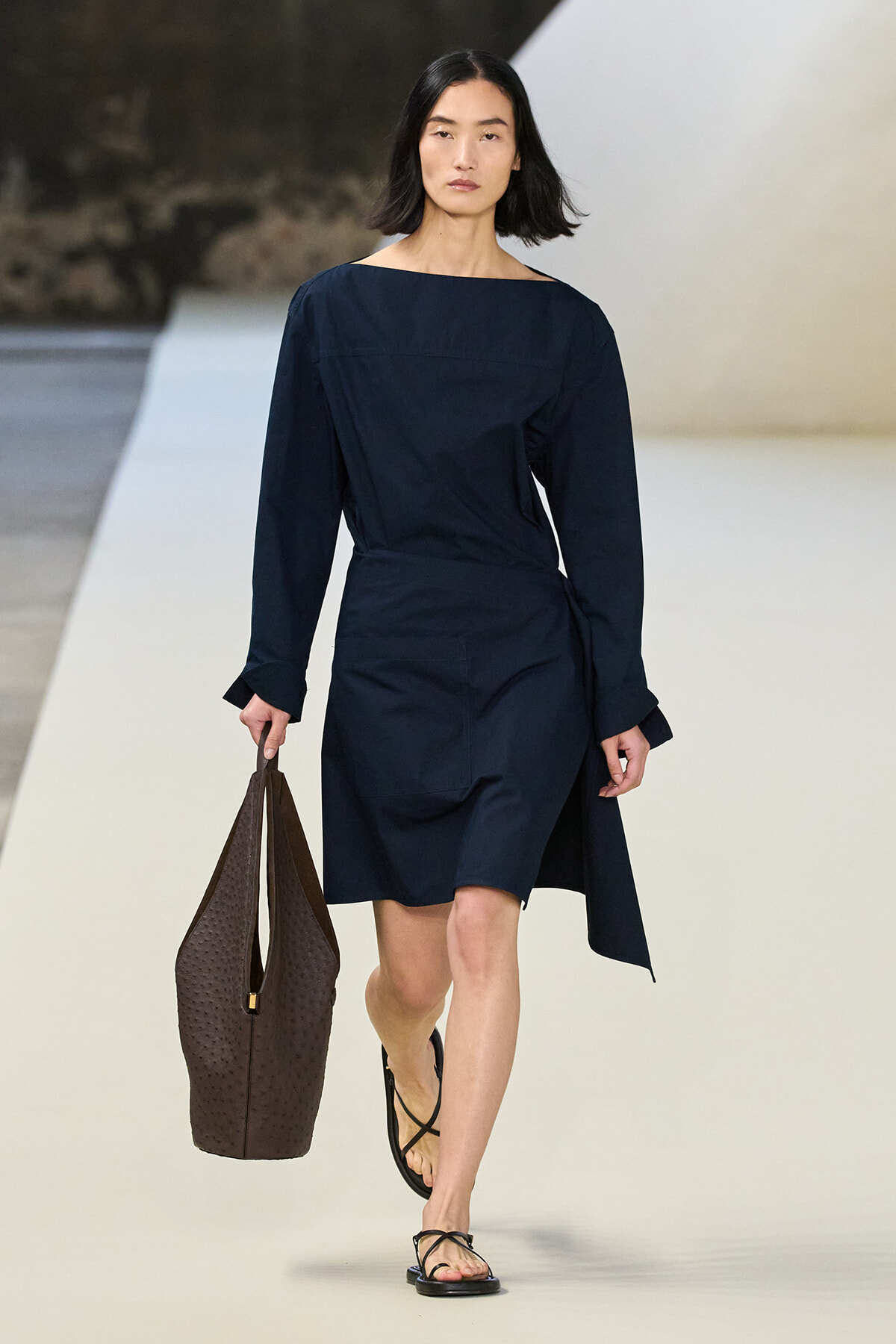 Model on a runway wearing a navy wrap dress with long sleeves, carrying a large brown textured tote and wearing black strappy sandals.