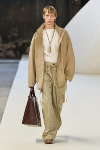 Male model walks a runway in a monochrome beige outfit: oversized tan jacket, matching cargo pants, white top, and a dark brown handbag.