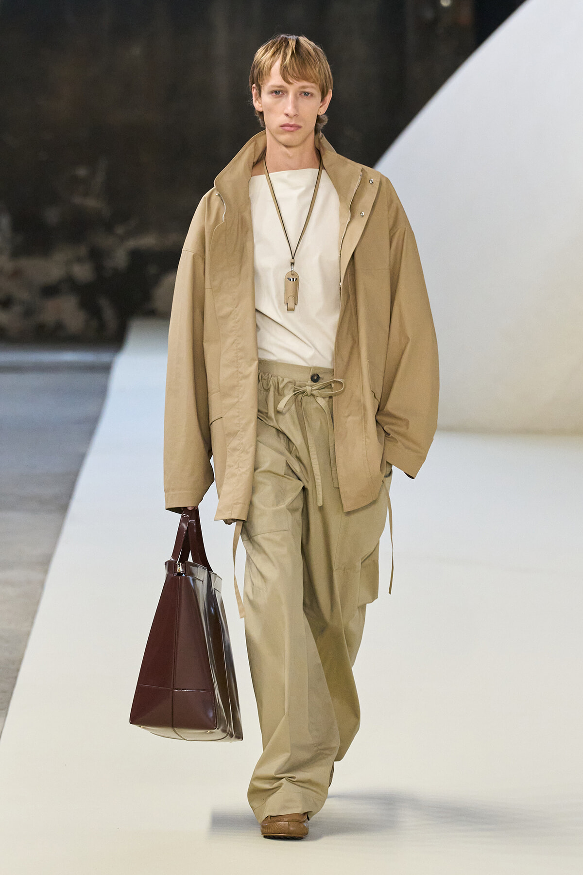 Male model walks a runway in a monochrome beige outfit: oversized tan jacket, matching cargo pants, white top, and a dark brown handbag.