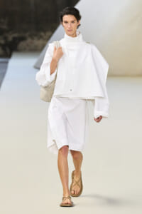 Model walking a white oversized cape-like top and knee-length white skirt on a fashion runway, carrying a beige woven bag and wearing tan sandals