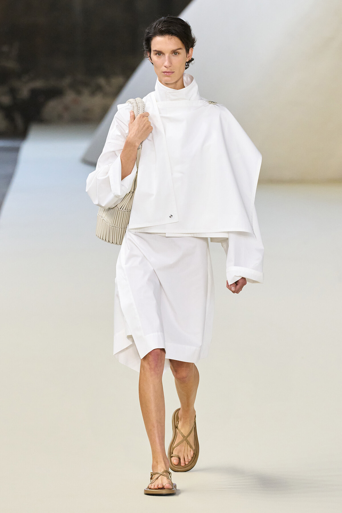 Model walking a white oversized cape-like top and knee-length white skirt on a fashion runway, carrying a beige woven bag and wearing tan sandals