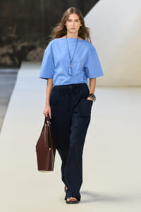 Model on a runway wearing a light blue, loose-fitting top and navy wide-leg pants, carrying a brown handbag and wearing a long necklace and bracelets.