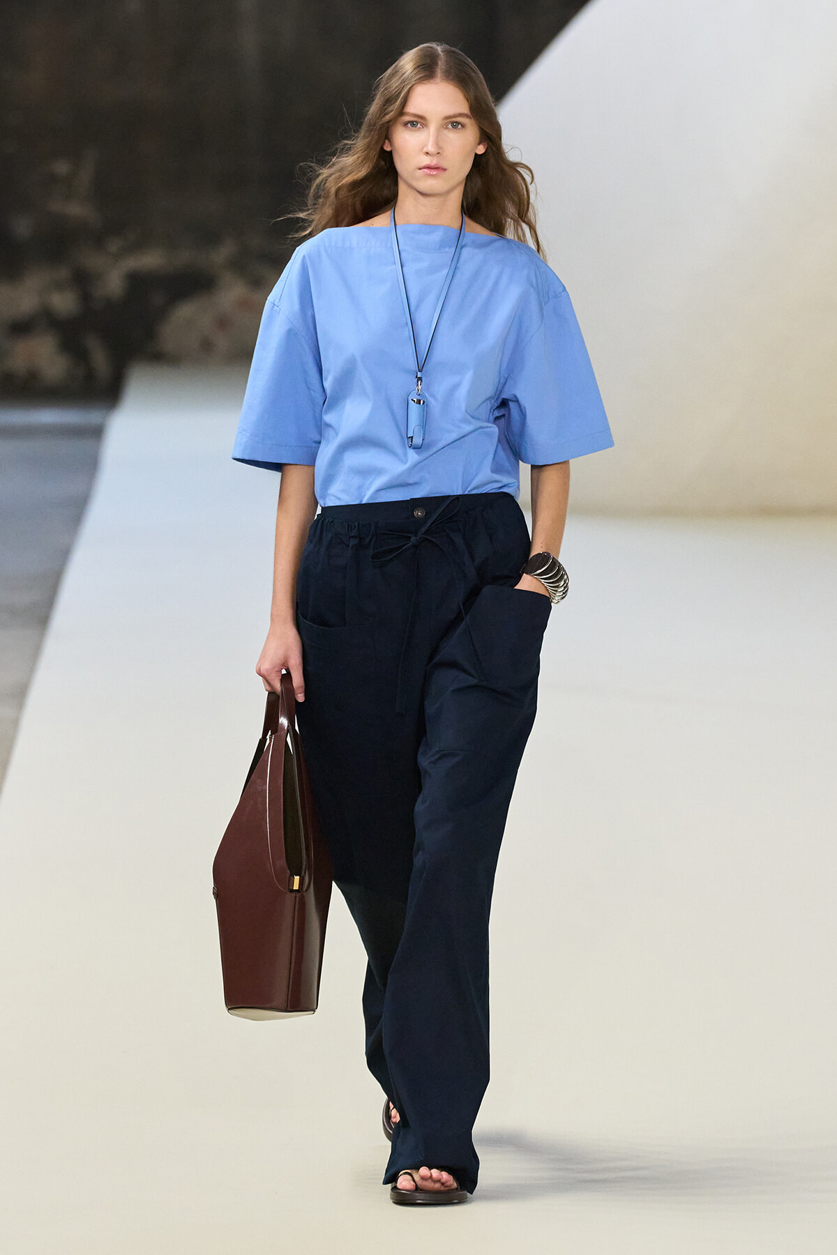 Model on a runway wearing a light blue, loose-fitting top and navy wide-leg pants, carrying a brown handbag and wearing a long necklace and bracelets.