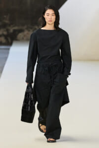 Model walking a runway in an all-black outfit: long-sleeve top with wide-legged pants, carrying a black woven tote and wearing flat sandals.