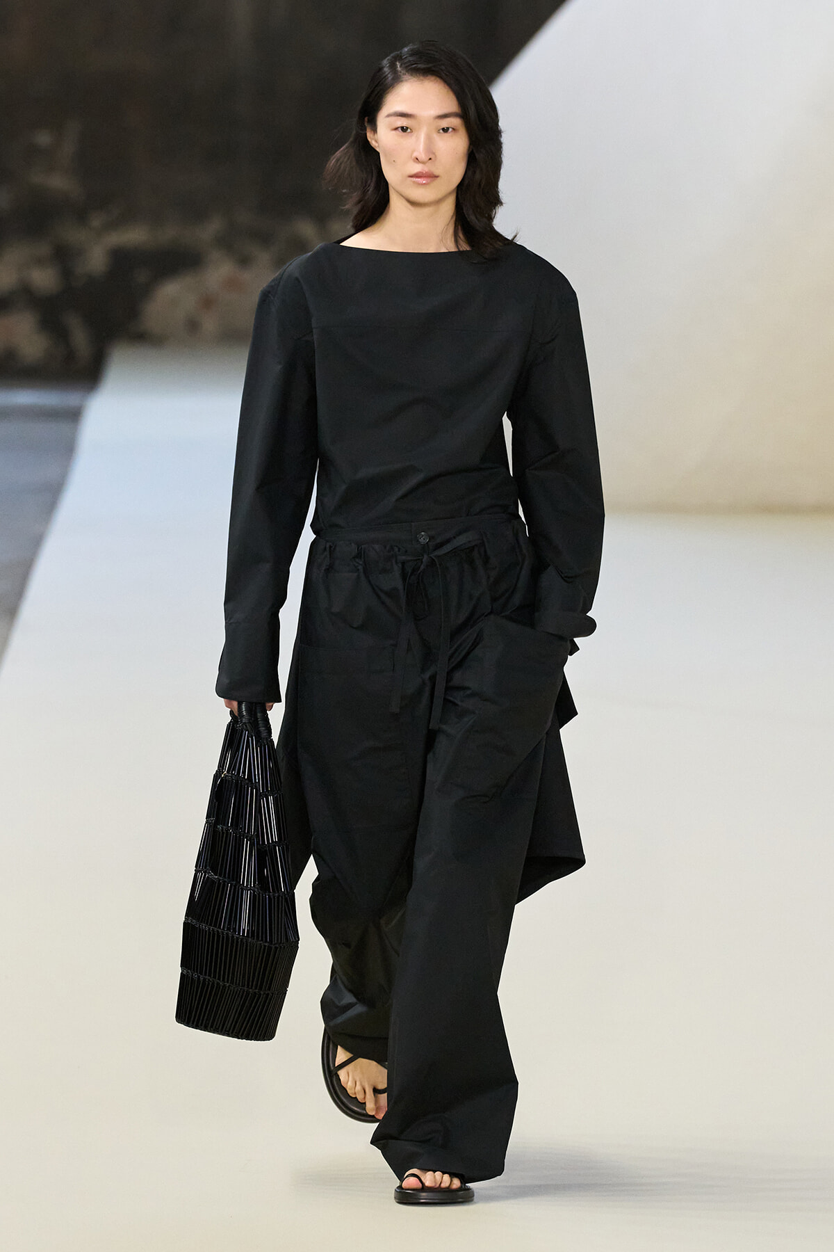 Model walking a runway in an all-black outfit: long-sleeve top with wide-legged pants, carrying a black woven tote and wearing flat sandals.