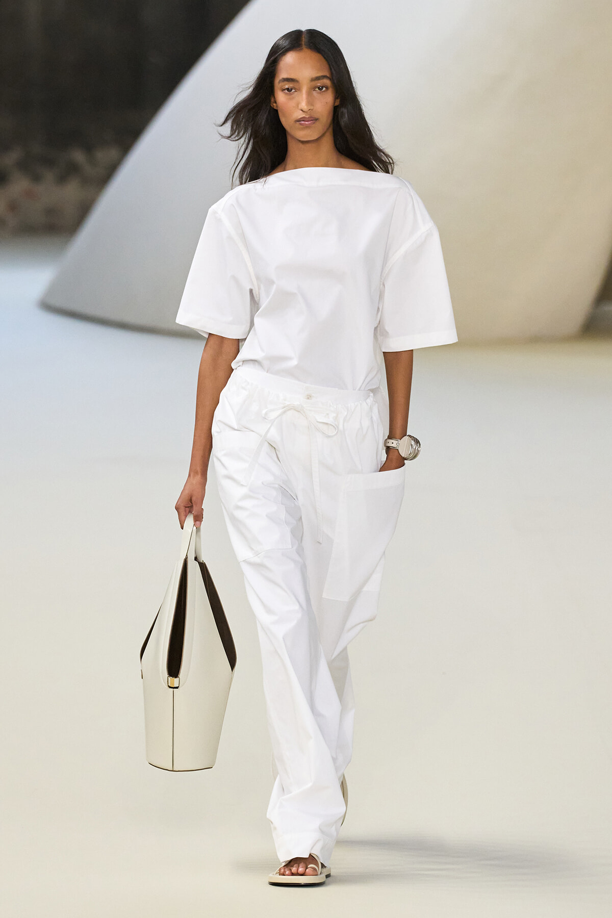 Model walking a runway in an all-white outfit: loose short-sleeve top, drawstring pants, beige sandals, and a cream tote bag.