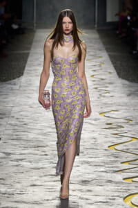 Slim model walking a runway in a lavender floral strapless dress with a matching choker and pink clutch.