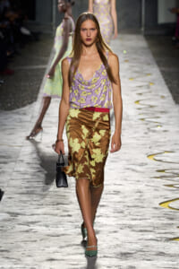 Runway model walking in a lilac floral draped top and brown satin skirt with yellow appliqué, carrying a small black handbag.