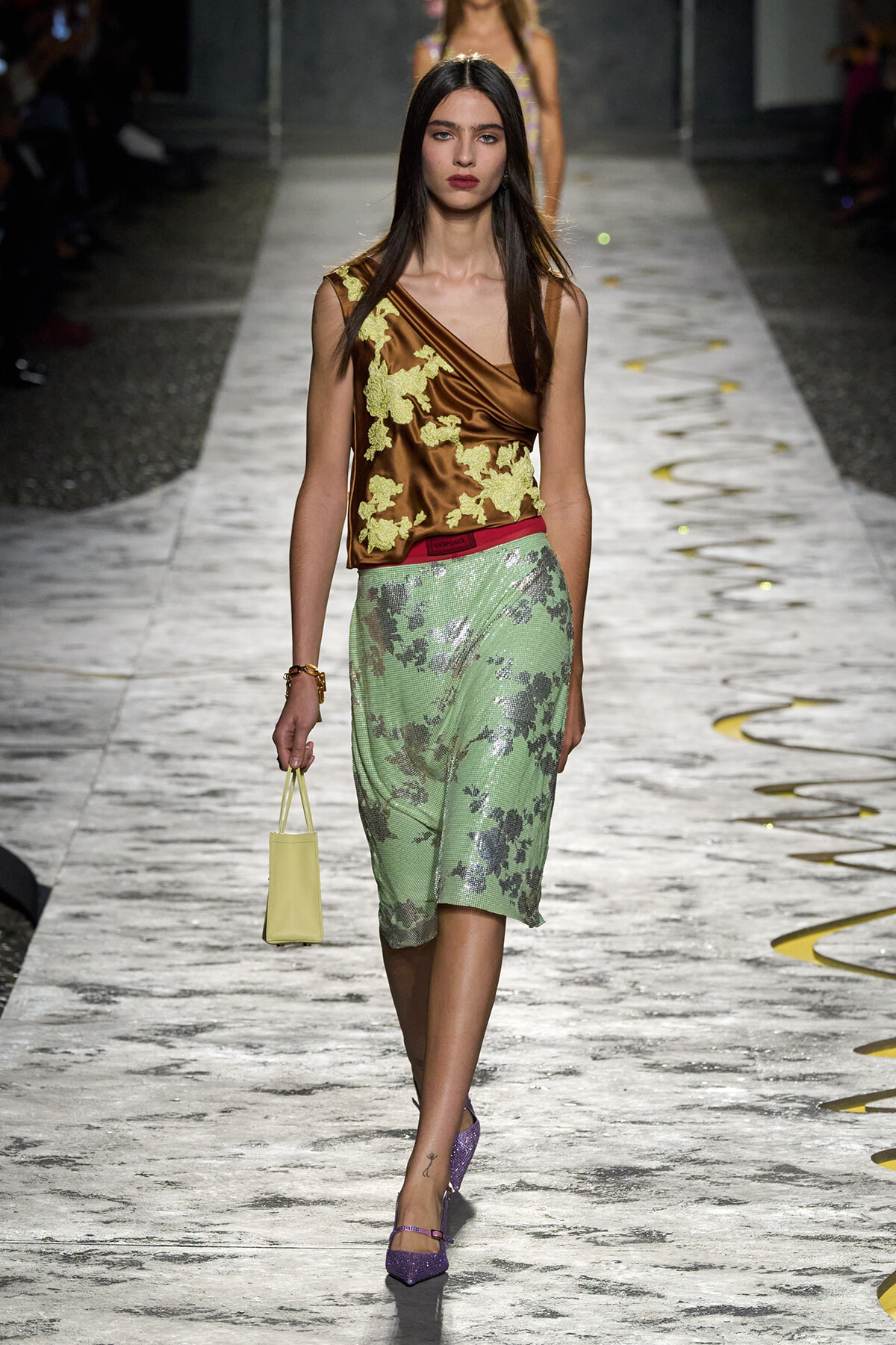 Runway model in bronze satin top with yellow floral embroidery and a mint-green floral skirt, carrying a small pale-yellow bag.