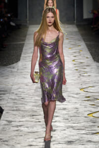 Model walks the runway in a shimmering lilac-gold metallic slip dress, holding a small clear clutch.