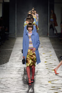 Model walks a fashion runway in a light blue coat over a zigzag top and metallic floral skirt with red tights and platform sandals.