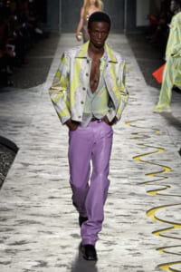 Male model walks a fashion runway wearing a pastel floral yellow jacket over a light shirt, lavender pants, and black boots, looking forward.