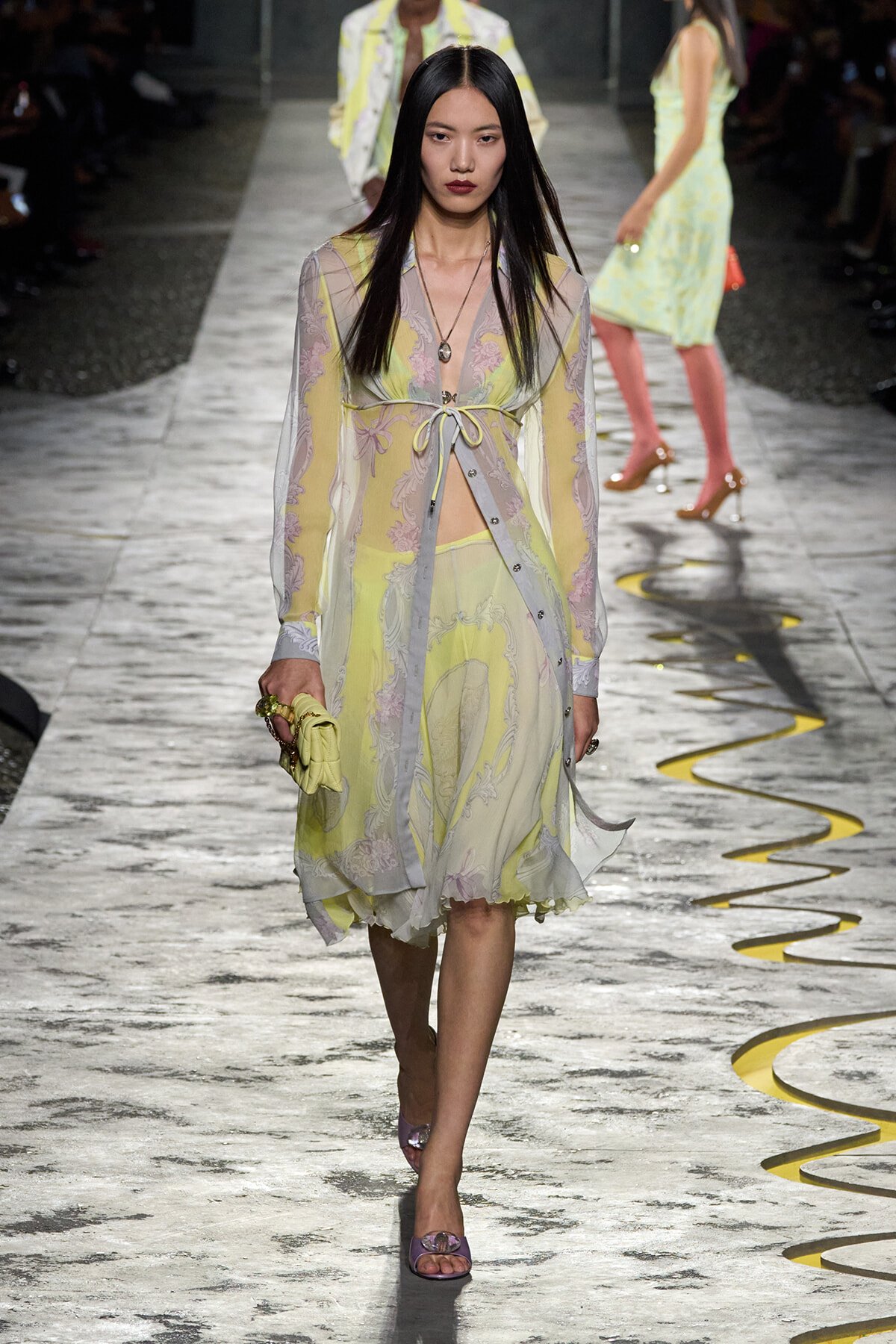 Model walking a fashion runway in a sheer pastel floral dress with a waist tie, holding a yellow clutch and wearing purple heels.