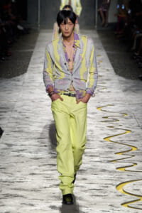 Male model walking a fashion show in a pastel yellow and lavender floral cardigan with matching trousers, hands in pockets, on a marble runway.