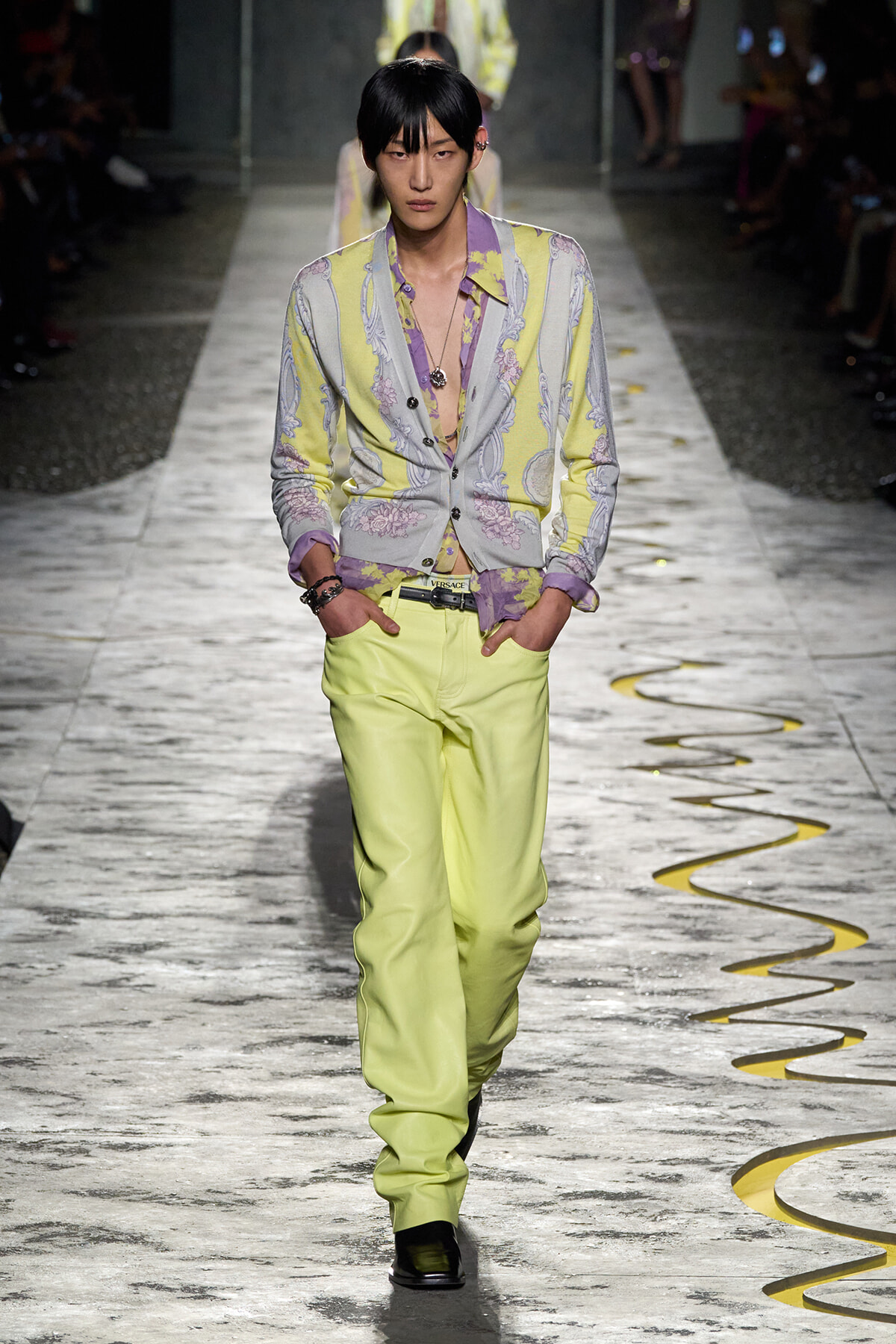 Male model walking a fashion show in a pastel yellow and lavender floral cardigan with matching trousers, hands in pockets, on a marble runway.