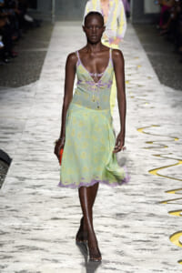 Model walks the runway in a pastel mint-green floral dress with lilac trim and a tied waist, during a fashion show on a marble catwalk.
