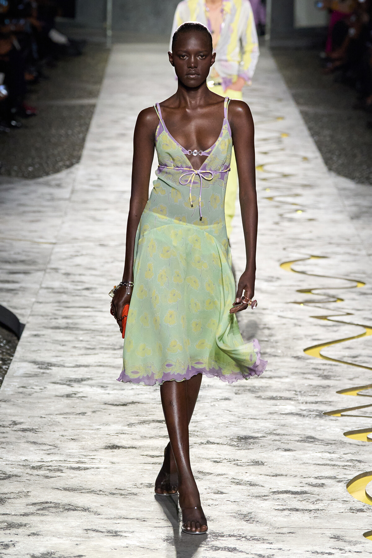 Model walks the runway in a pastel mint-green floral dress with lilac trim and a tied waist, during a fashion show on a marble catwalk.