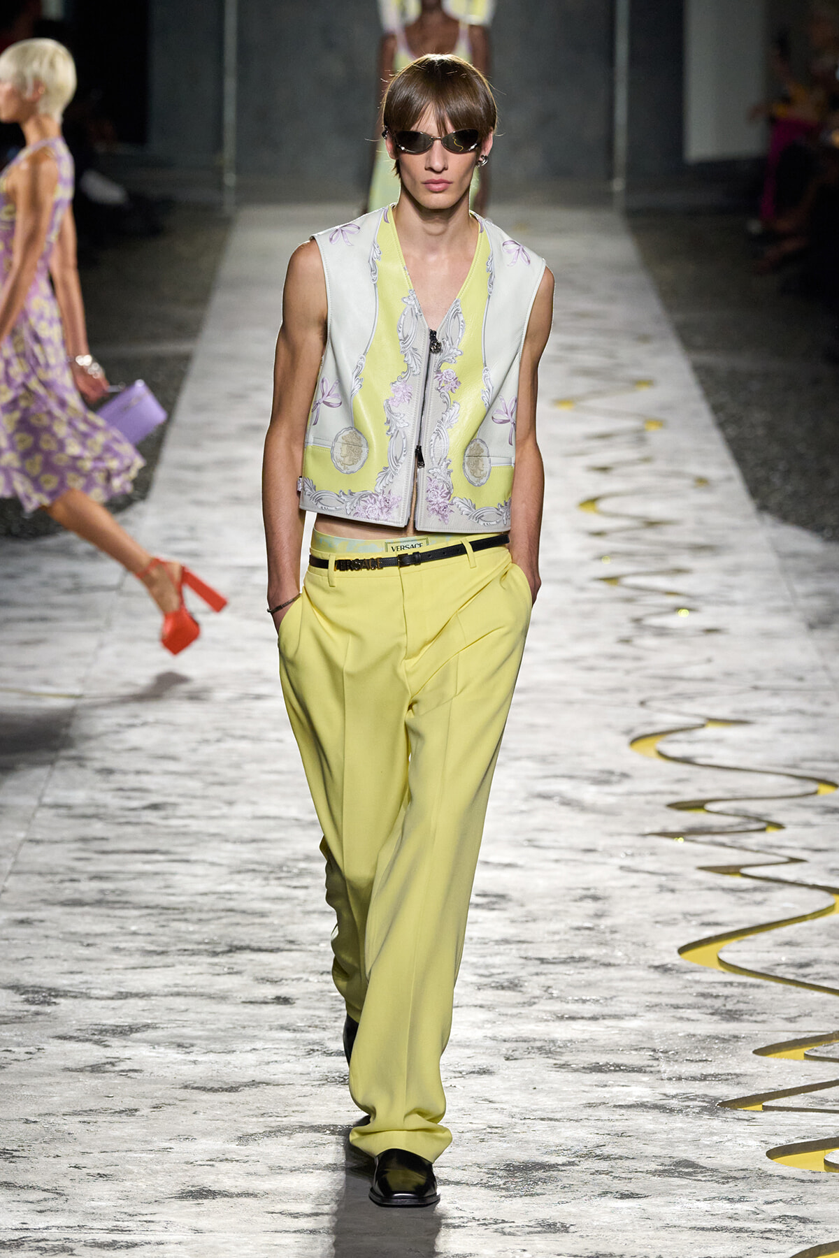 Model walks a fashion runway in pale embroidered vest and bright yellow wide-leg trousers, wearing sunglasses and black shoes.