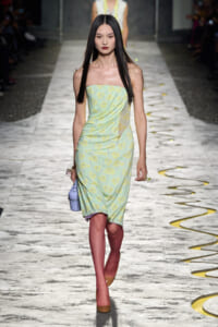 Model walking a fashion runway in a strapless pastel green floral dress, long black hair, brown heels, and a light purple handbag.