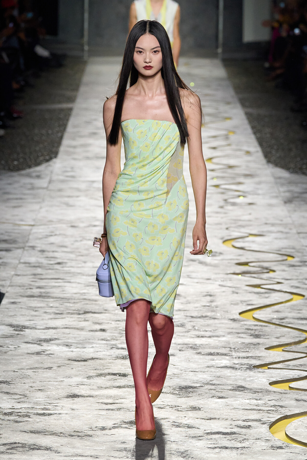 Model walking a fashion runway in a strapless pastel green floral dress, long black hair, brown heels, and a light purple handbag.