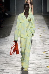 Model walking a runway in a pale green tie-dye suit, paired with a bright red handbag.