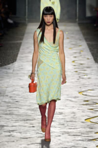 Model walking a runway in a pastel green floral dress, holding a small orange cylindrical handbag.