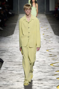 Model walking a runway in a pale yellow, loose-fit blazer and matching wide-leg pants with large pockets and a single chest pocket.