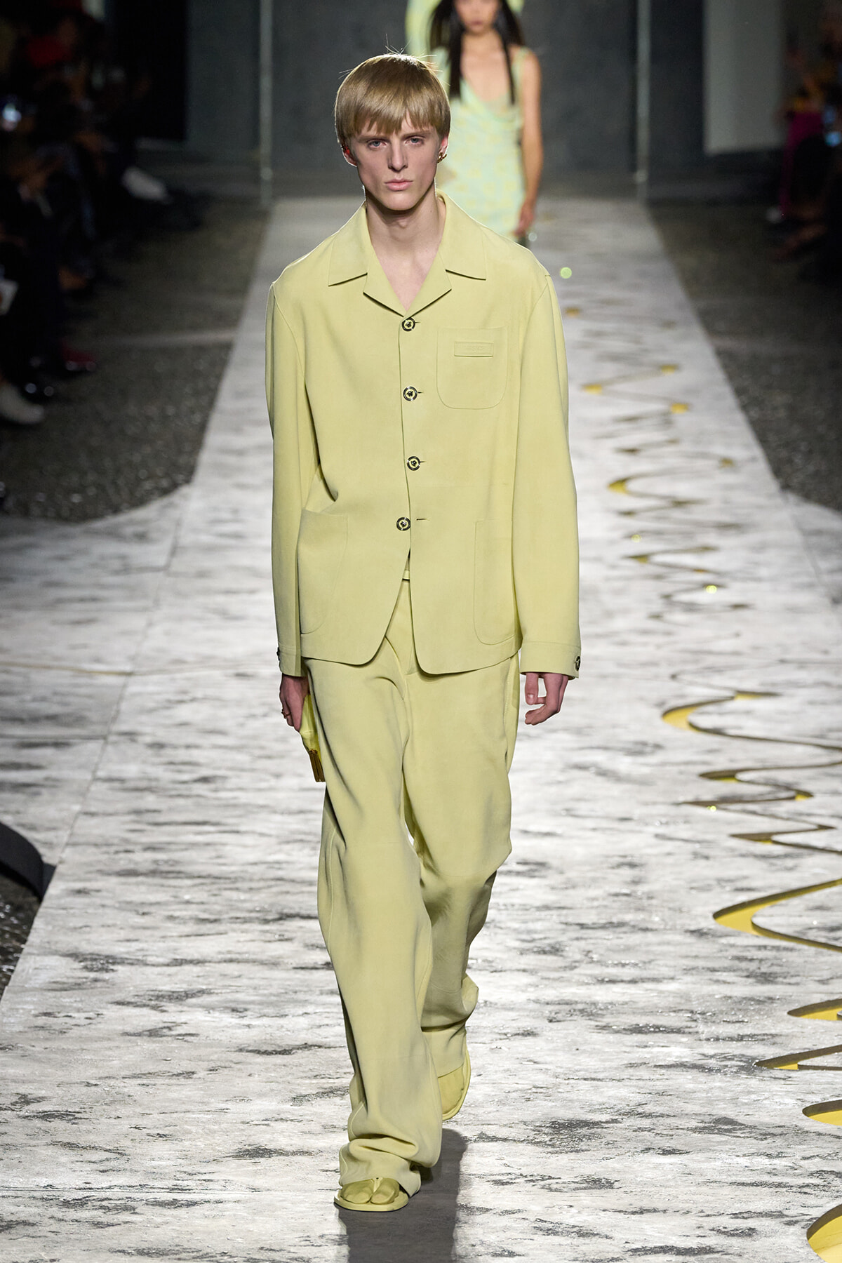 Model walking a runway in a pale yellow, loose-fit blazer and matching wide-leg pants with large pockets and a single chest pocket.