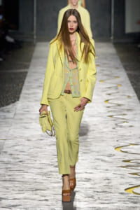 Model walks the runway in a pale yellow blazer and matching trousers, paired with a light green floral top and a small quilted yellow handbag with a chain strap.