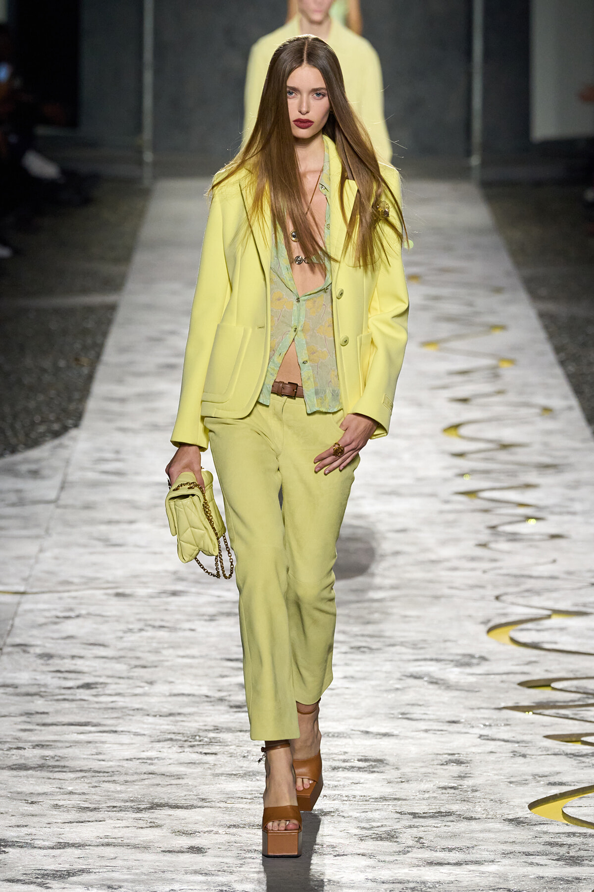 Model walks the runway in a pale yellow blazer and matching trousers, paired with a light green floral top and a small quilted yellow handbag with a chain strap.