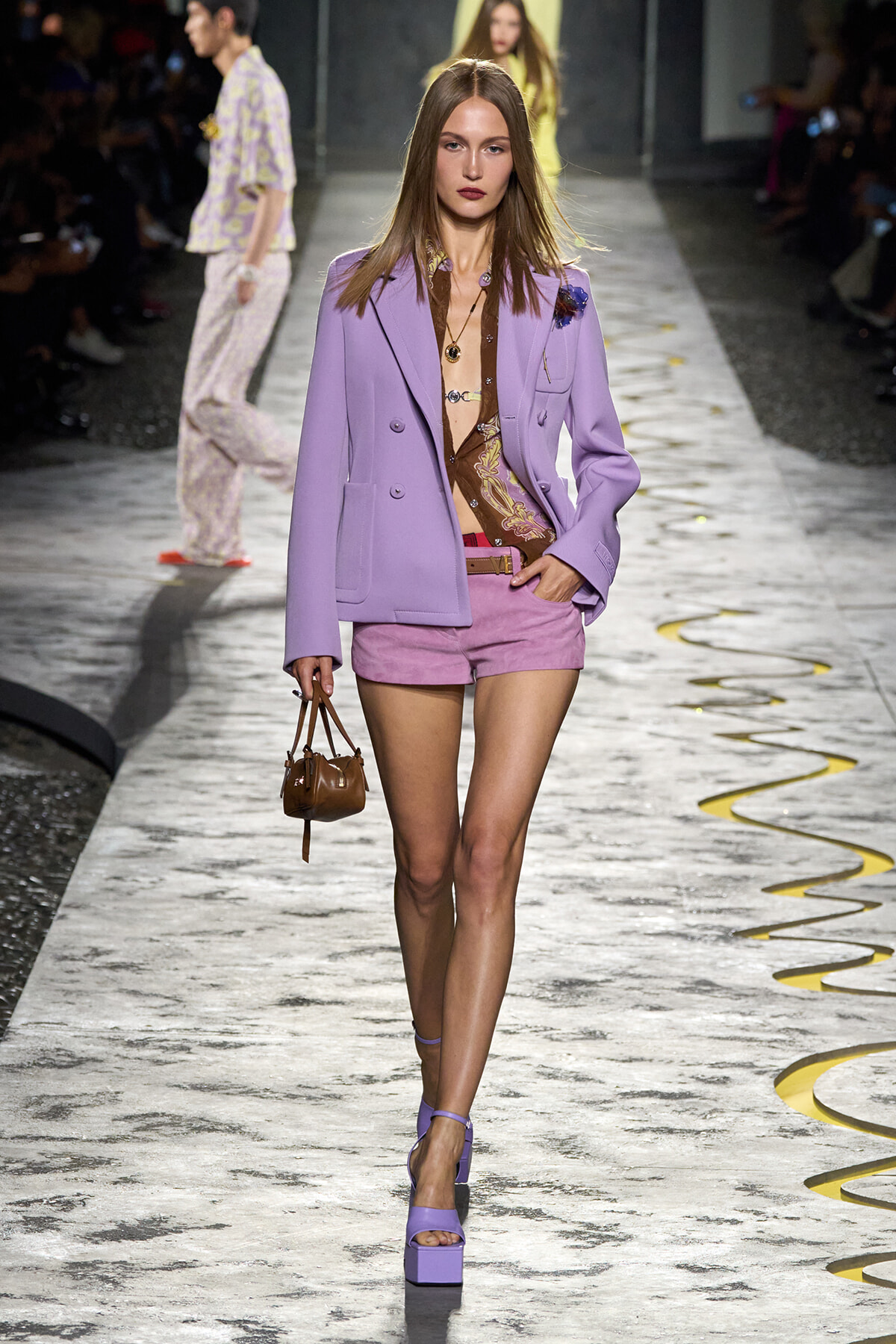 Model walks the runway in a lilac blazer, pink shorts, and brown printed top, carrying a small brown handbag with purple platform sandals.