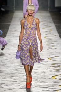 Model walking the runway in a lavender floral midi dress with a front slit, paired with a small lavender handbag and orange platform sandals.