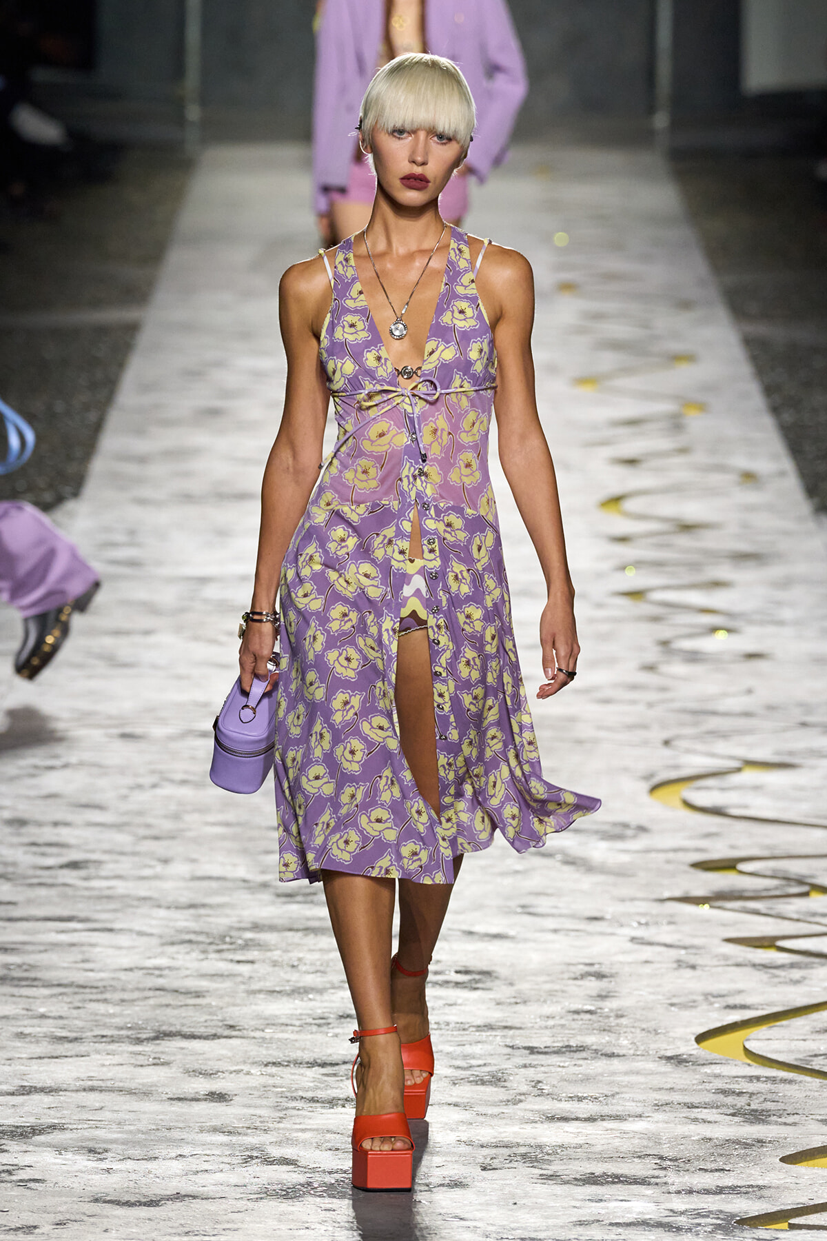 Model walking the runway in a lavender floral midi dress with a front slit, paired with a small lavender handbag and orange platform sandals.