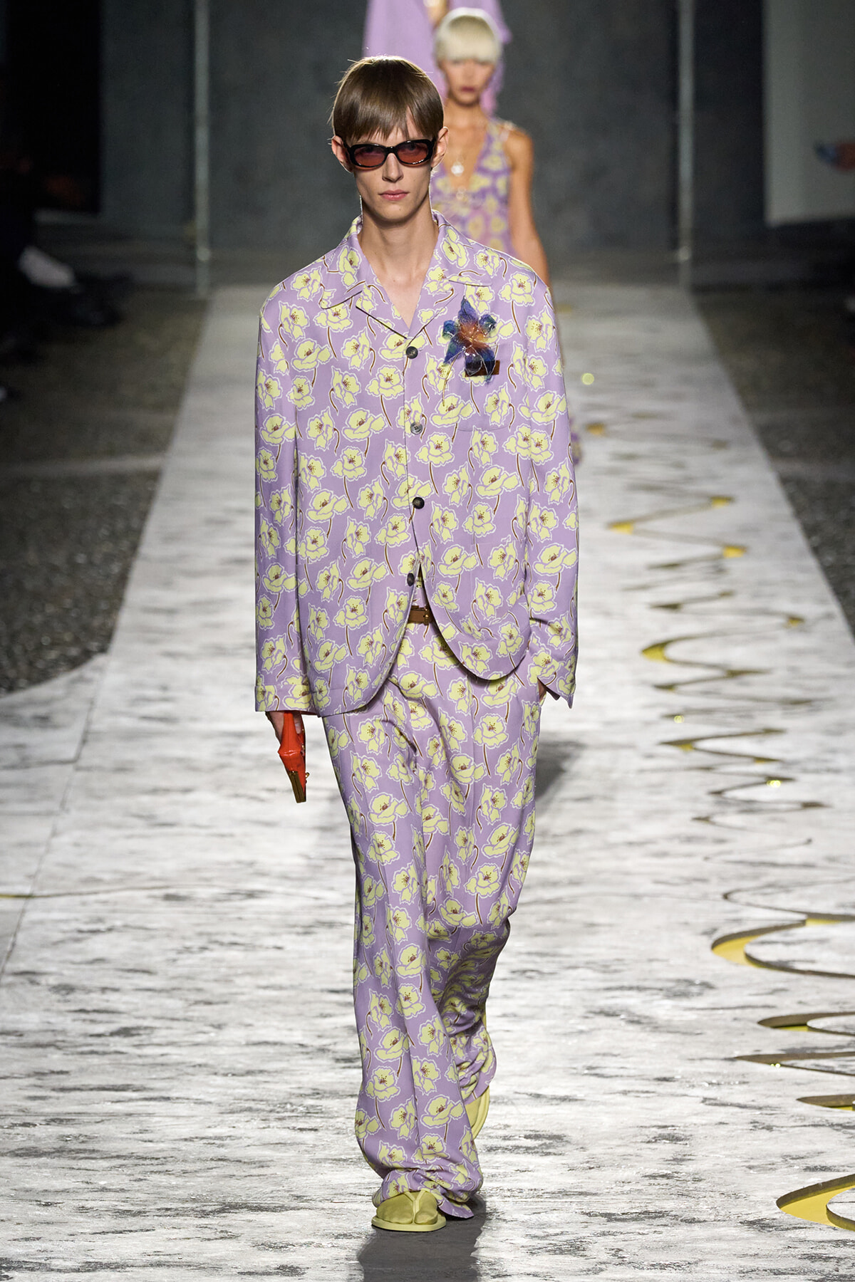 Model walking the runway in a lavender floral suit with matching pants and oversized sunglasses.