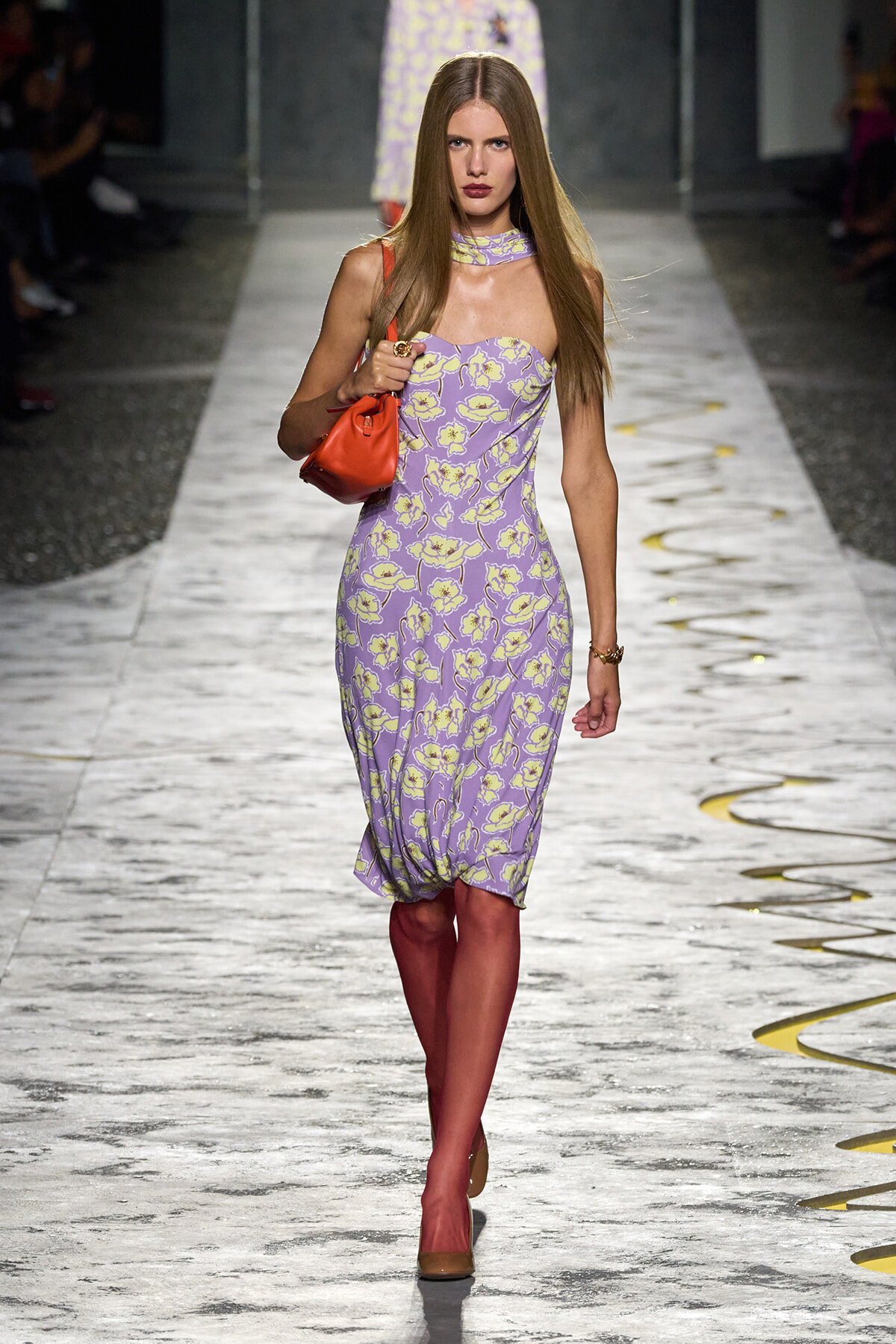 Model walking a fashion runway in a lavender yellow floral dress with an orange handbag and brown heels.
