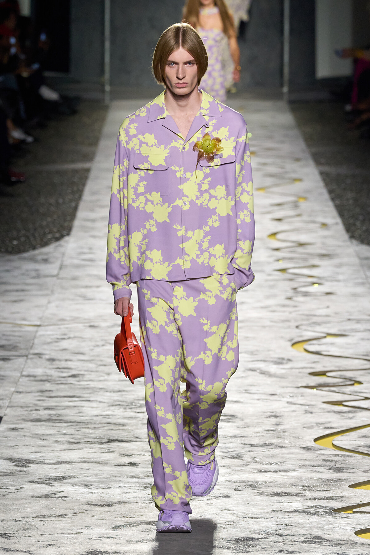 Model on a runway wearing a lavender suit with pale yellow floral pattern, carrying a small red handbag.
