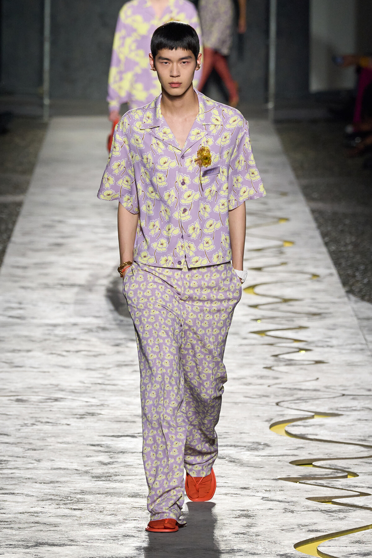 Male model walking a runway in lavender floral pajama-style suit with yellow flowers and orange footwear, wearing a yellow flower brooch on the chest.