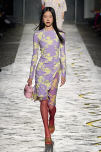 Model walks a runway in a lavender dress with yellow floral print, carrying a pink handbag and wearing tan sandals.