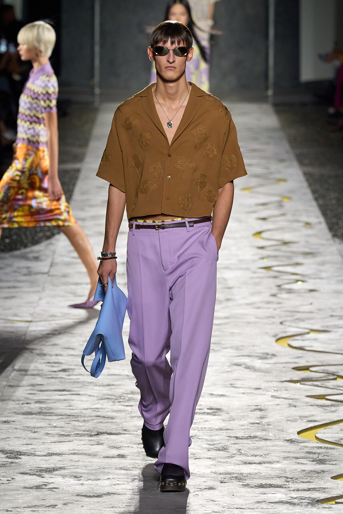 Male model on a runway wearing a brown floral short-sleeve shirt with lavender trousers, black sunglasses, and a light blue bag in hand; minimal jewelry visible as he walks.