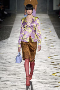 Model walking runway in lavender satin blouse with yellow floral pattern, brown lace-trimmed skirt, pink tights, red sandals, and a light-blue handbag; black bob hairstyle and bold lipstick.