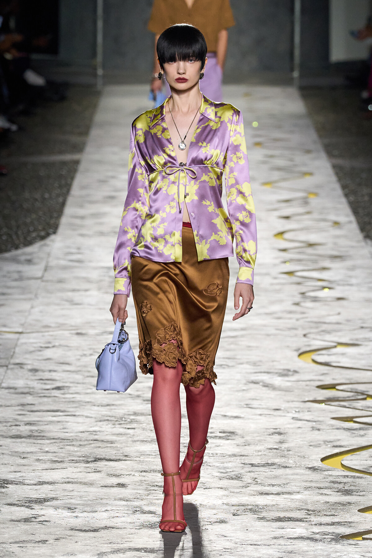 Model walking runway in lavender satin blouse with yellow floral pattern, brown lace-trimmed skirt, pink tights, red sandals, and a light-blue handbag; black bob hairstyle and bold lipstick.