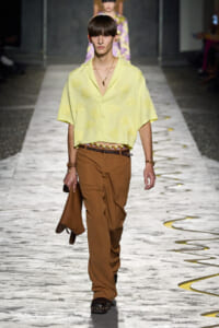 Model walks the runway in a pale yellow embroidered blouse and brown trousers, holding a brown jacket.