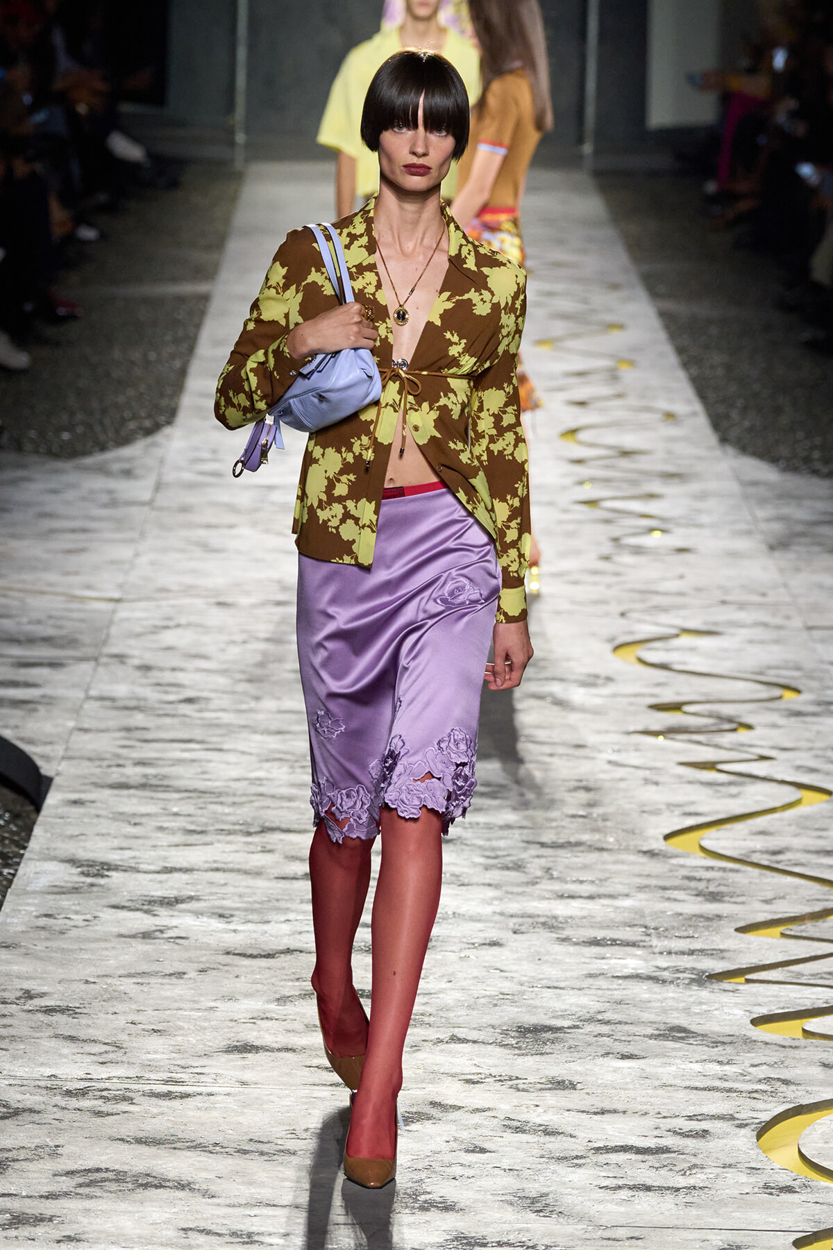 Model walking a fashion runway in a brown and lime floral blazer over lavender satin skirt with lace hem, carrying a lavender bag.