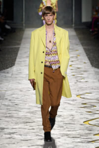 Model on a runway wearing a pale yellow overcoat, a purple patterned shirt, brown trousers, and black boots with a pendant necklace and clutch in hand.