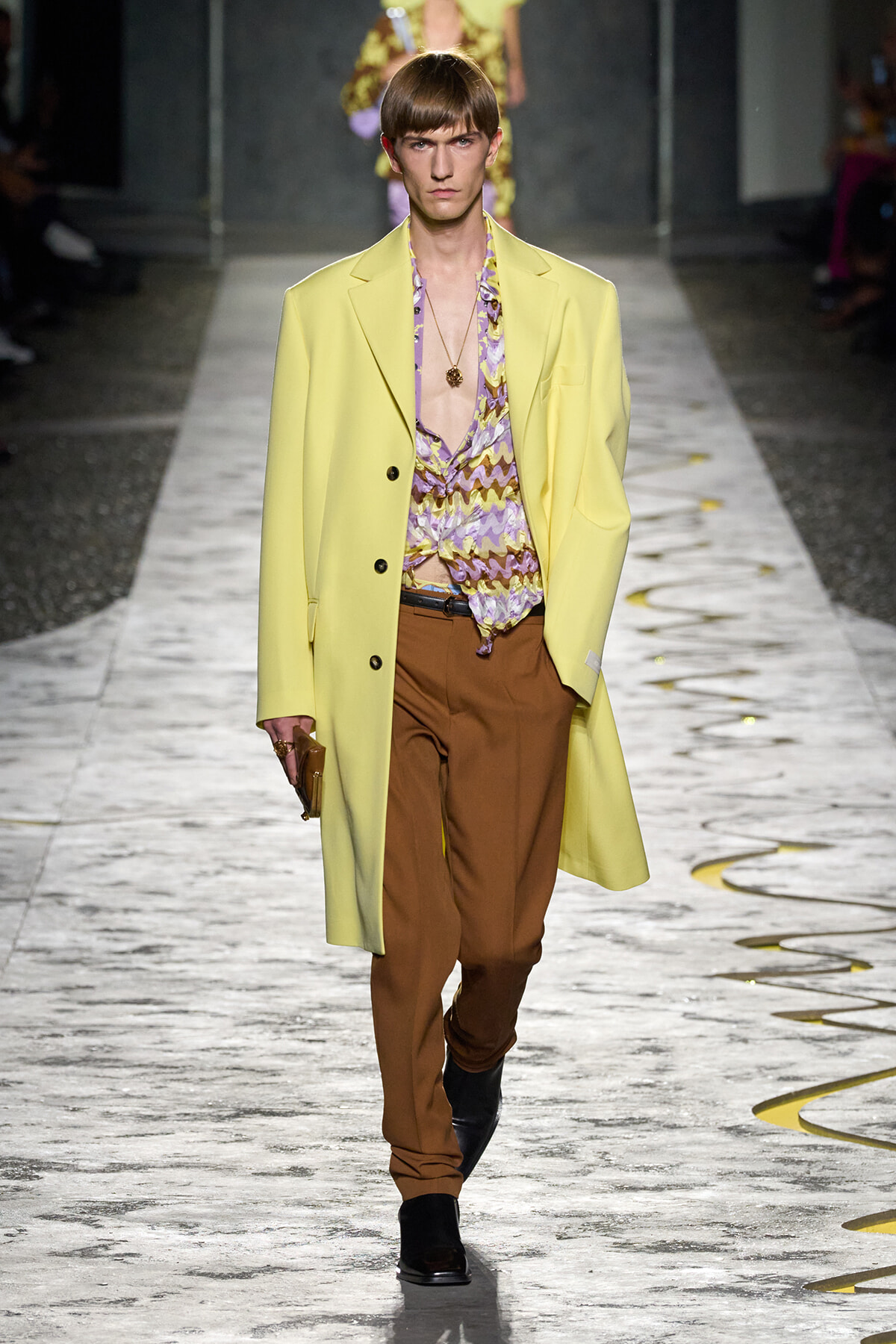 Model on a runway wearing a pale yellow overcoat, a purple patterned shirt, brown trousers, and black boots with a pendant necklace and clutch in hand.